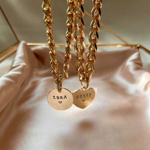 May include: Two gold-toned chain necklaces featuring flat, interlocking links. One necklace has a circular pendant engraved with the name LUNA and a small heart. The other necklace has a heart-shaped pendant engraved with the name RILEY and a small heart.