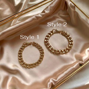 May include: Two gold-toned metal chain bracelets with lobster claw clasps. The bracelet labeled Style 1 features a smaller link design, while the bracelet labeled Style 2 features a larger, thicker link design. Both items are metallic gold.
