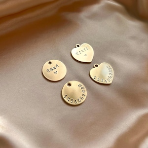 May include: Four gold-toned metal charms with circular and heart shapes. Two charms are circular, one stamped with the name LUNA and a heart, the other with the numbers 1234567890. Two are heart-shaped, one stamped RILEY and a heart, the other 1234567890.