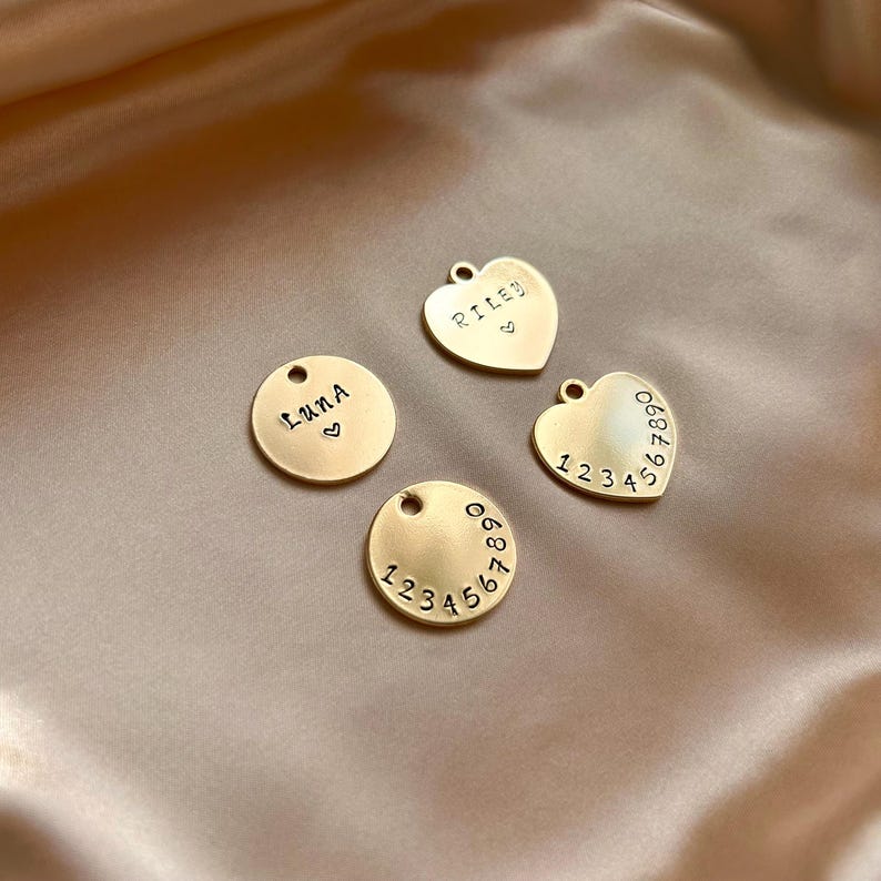 May include: Four gold-toned metal charms with circular and heart shapes. Two charms are circular, one stamped with the name LUNA and a heart, the other with the numbers 1234567890. Two are heart-shaped, one stamped RILEY and a heart, the other 1234567890.