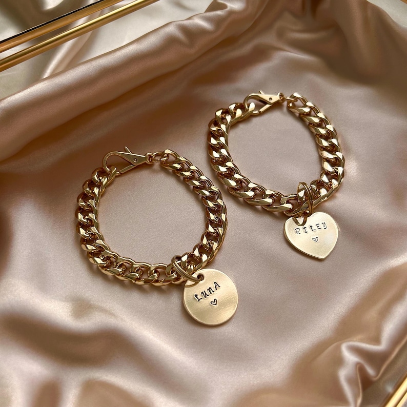 May include: Two gold-toned metal chain bracelets featuring lobster claw clasps. One bracelet has a round pendant engraved with the name Luna and a small heart, while the other has a heart-shaped pendant engraved with the name Riley and a heart.