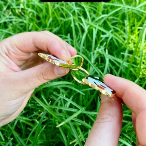 K&ouml;nnte beinhalten: Nahaufnahme eines goldfarbenen Schl&uuml;sselanh&auml;ngers aus Metall mit einem roten, wei&szlig;en und schwarzen Emaille-Design. Der Schl&uuml;sselanh&auml;nger ist 2,2 mm dick.  THICKNESS ist in gelber Schrift oben im Bild zu sehen.