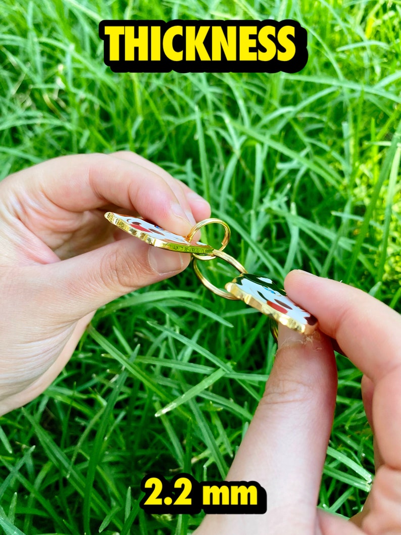 K&ouml;nnte beinhalten: Nahaufnahme eines goldfarbenen Schl&uuml;sselanh&auml;ngers aus Metall mit einem roten, wei&szlig;en und schwarzen Emaille-Design. Der Schl&uuml;sselanh&auml;nger ist 2,2 mm dick.  THICKNESS ist in gelber Schrift oben im Bild zu sehen.