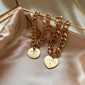 May include: Two gold-toned chain bracelets with thick, interlocking links. One bracelet features a round pendant engraved with the name Luna and a small heart. The other bracelet features a heart-shaped pendant engraved with the name Riley and a small heart.
