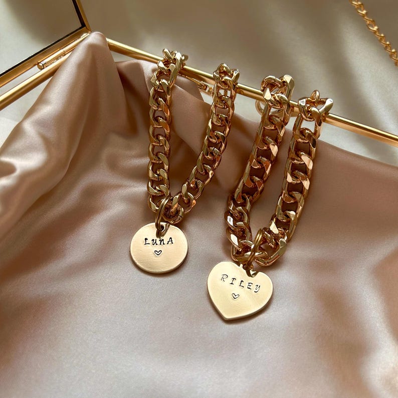 May include: Two gold-toned chain bracelets with thick, interlocking links. One bracelet features a round pendant engraved with the name Luna and a small heart. The other bracelet features a heart-shaped pendant engraved with the name Riley and a small heart.