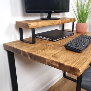 May include: A wooden desk with black metal legs and a shelf for a monitor. A laptop, keyboard, and mouse are on the desk. A potted plant and a black metal wine rack are in the background.