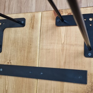 May include: The underside of a wooden table with black metal hairpin legs. The legs are attached to the table with screws. There is a black metal bar across the bottom of the table.