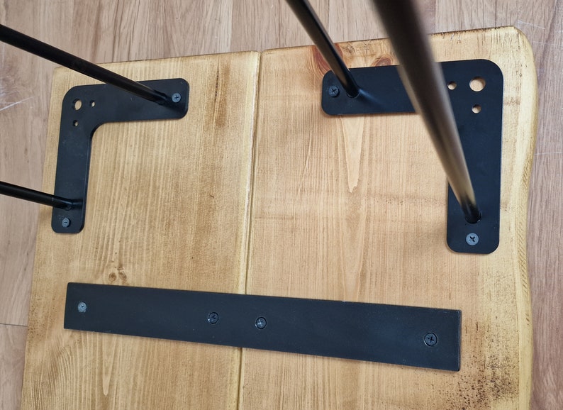 May include: The underside of a wooden table with black metal hairpin legs. The legs are attached to the table with screws. There is a black metal bar across the bottom of the table.