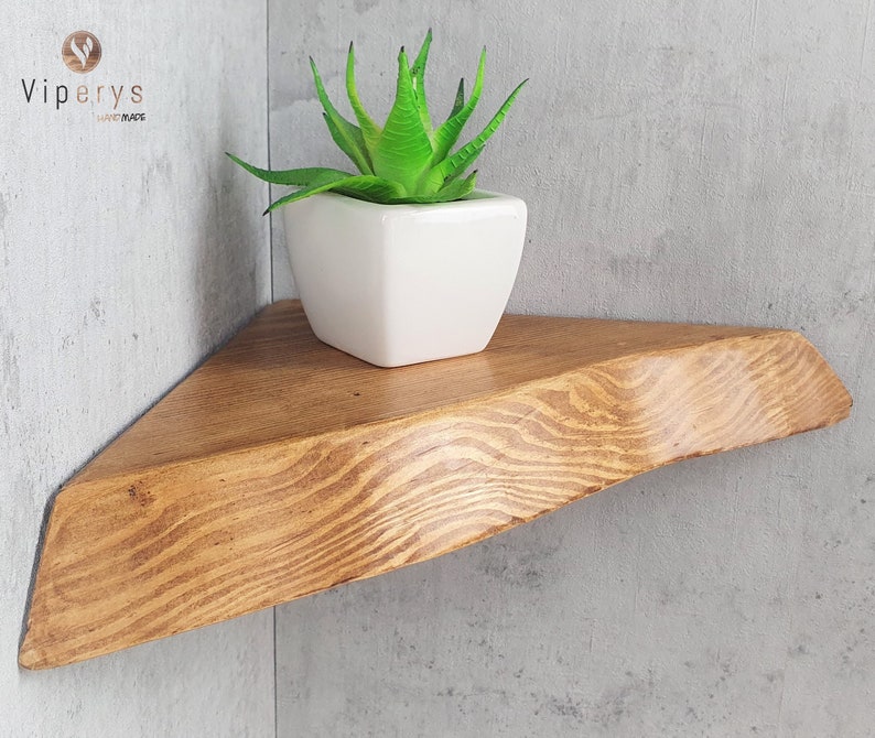May include: A triangular wooden shelf with a natural wood finish. The shelf is mounted on a corner wall and has a small succulent plant in a white ceramic pot on top.