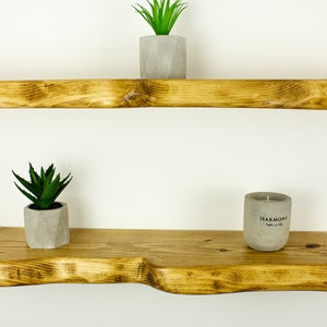 May include: Two wooden shelves with a rustic, natural finish. The top shelf is straight, while the bottom shelf has a curved edge. The shelves are decorated with small potted plants and a candle.