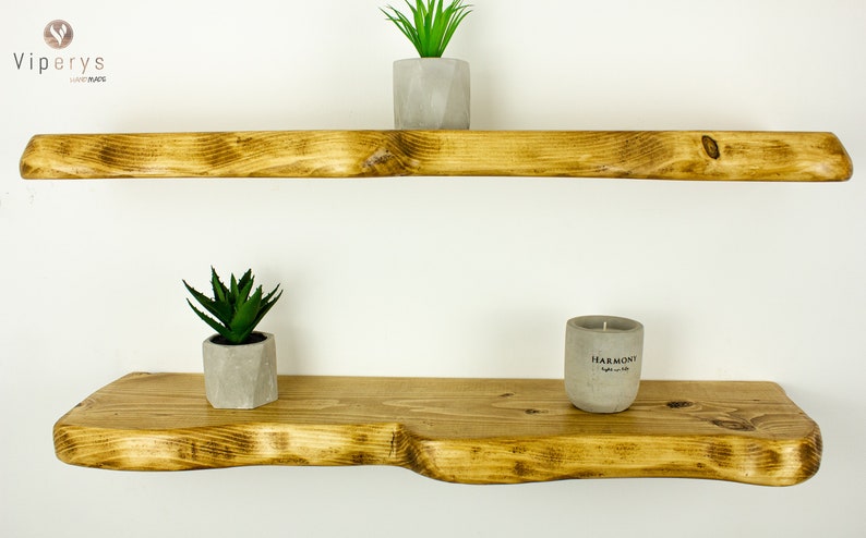 May include: Two wooden shelves with a rustic, natural finish. The top shelf is straight, while the bottom shelf has a curved edge. The shelves are decorated with small potted plants and a candle.