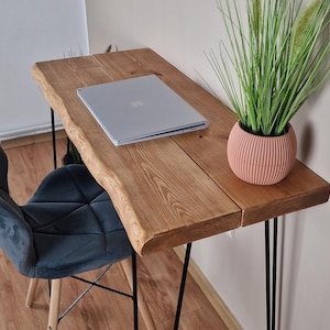May include: A wooden desk with a live edge top and black hairpin legs. A laptop computer is on the desk, and a potted plant is on the right side of the desk. A dark blue velvet chair is in the foreground.