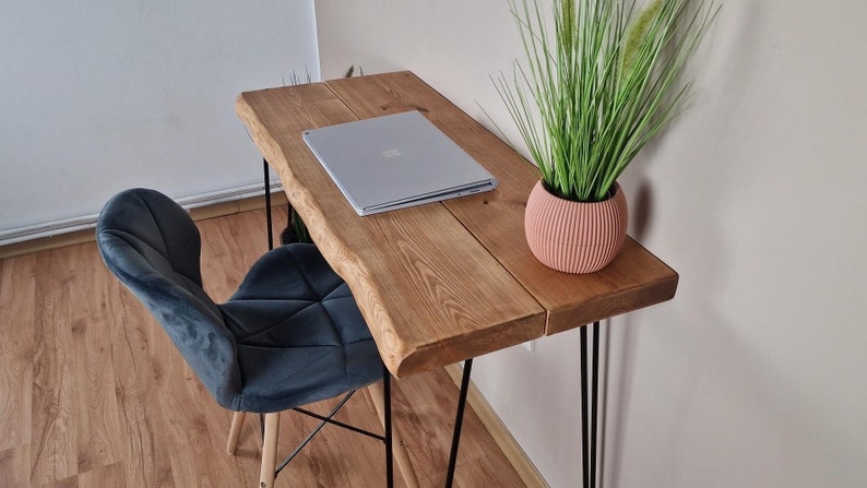 May include: A wooden desk with a live edge top and black hairpin legs. A laptop computer is on the desk, and a potted plant is on the right side of the desk. A dark blue velvet chair is in the foreground.