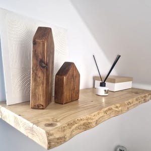 May include: A rustic wooden shelf with a live edge, displaying decorative items. Two wooden house-shaped sculptures and a tall rectangular wooden piece stand on the shelf. A white canvas with a textured design and a diffuser with black reeds complete the display.