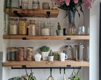 May include: Wooden shelves with black metal brackets and hooks, filled with various items, including jars of spices, succulents, and a vase of pink lilies.