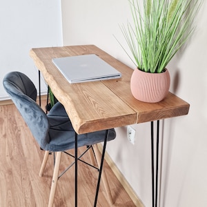 May include: A wooden desk with a live edge top and black hairpin legs. A laptop computer and a potted plant are on the desk. A gray velvet chair is in front of the desk.