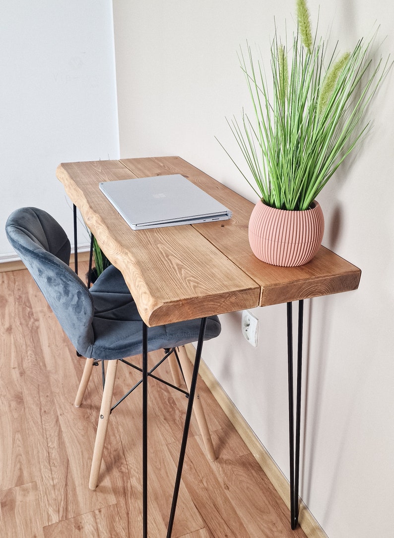 May include: A wooden desk with a live edge top and black hairpin legs. A laptop computer and a potted plant are on the desk. A gray velvet chair is in front of the desk.