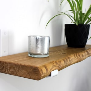 Könnte beinhalten: Ein rustikales Holzregal mit natürlicher Kante, getragen von weißen Halterungen, an einer weißen Wand befestigt. Ein silberner Glaskerzenhalter und ein schwarzer geometrischer Pflanztopf mit einer grünen Pflanze stehen auf dem Regal. Der Kerzenhalter trägt den Text "Jasmine".