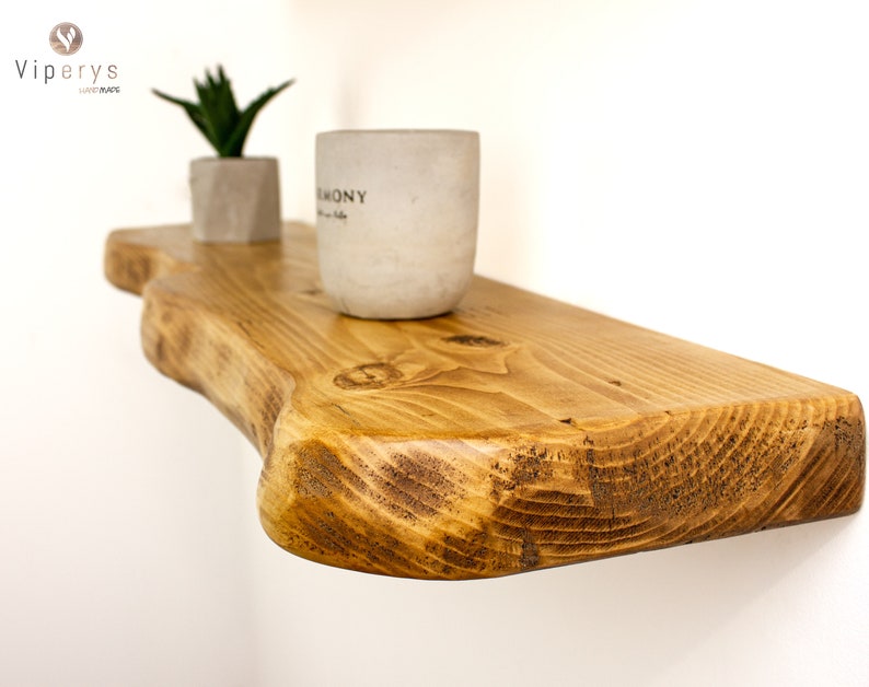 May include: A rustic wooden shelf with a natural, live edge design. The shelf is stained a warm brown color and has a smooth, polished finish. Two small concrete planters with plants are sitting on the shelf.