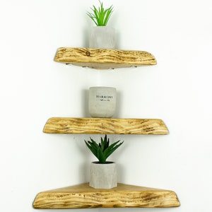 May include: Three wooden corner shelves with a rustic finish, stacked on a white wall. The top shelf has a small potted succulent plant, the middle shelf has a small white candle, and the bottom shelf has a small potted succulent plant.