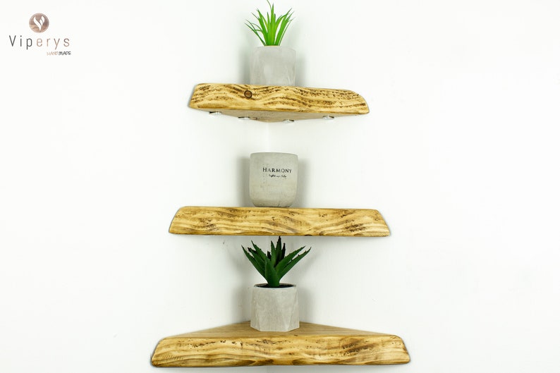 May include: Three wooden corner shelves with a rustic finish, stacked on a white wall. The top shelf has a small potted succulent plant, the middle shelf has a small white candle, and the bottom shelf has a small potted succulent plant.