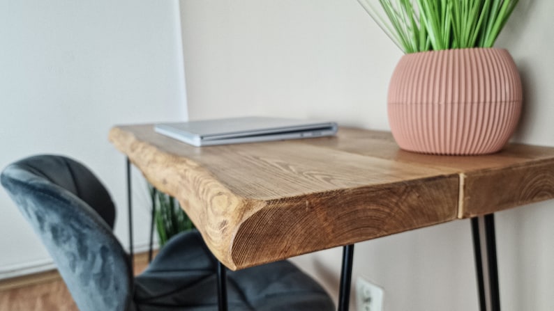 May include: A wooden desk with a live edge top and black hairpin legs. A laptop computer is on the desk, and a pink planter with green plants is on the right side of the desk.