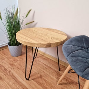 May include: A round wooden side table with a natural wood grain finish. The table has black hairpin legs. The table is approximately 18 inches in diameter and 18 inches tall. A gray chair and a potted plant are in the background.
