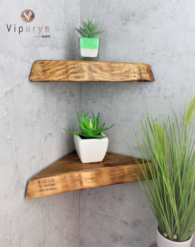 May include: Two wooden corner shelves with a rustic finish. The shelves are triangular and have a natural wood grain. The top shelf has a small green plant in a clear glass container. The bottom shelf has a small green plant in a white ceramic pot. The shelves are mounted on a gray concrete wall.