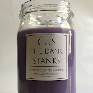 May include: A purple coffee scented candle in a glass jar with a white label that reads "CUS THE DANK STANKS COFFEE SCENTED CANDLE HANDMADE IN MILWAUKEE, WI CRUDE CANDLES".