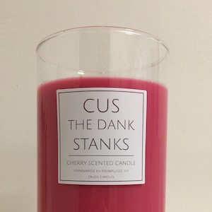 May include: A clear glass jar candle with a pink cherry scented wax. The label reads "CUS THE DANK STANKS" and "CHERRY SCENTED CANDLE" with the text "HANDMADE IN MILWAUKEE, WI" and "CRUDE CANDLES" below.