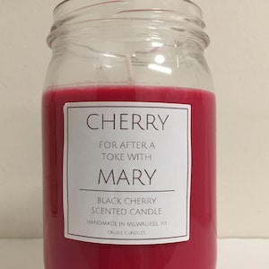 May include: A red glass jar candle with a white label that reads "CHERRY FOR AFTER A TOKE WITH MARY BLACK CHERRY SCENTED CANDLE HANDMADE IN MILWAUKEE, WI CRUDE CANDLES".