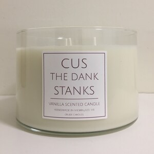 May include: A clear glass candle jar with a white label that reads "CUS THE DANK STANKS" in brown text. The label also says "Vanilla Scented Candle" and "Handmade in Milwaukee, WI" in smaller brown text.