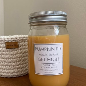 Può includere: Una candela in vetro in un barattolo con un coperchio argentato e un'etichetta bianca che recita "Pumpkin Pie For After You Get High Pumpkin Pie Scented Candle Handmade in Milwaukee, WI Crude Candles". La candela è di colore arancio chiaro e si trova su una superficie di legno.