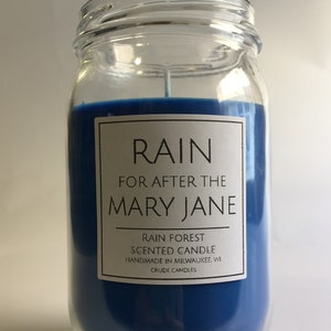Puede incluir: Una vela perfumada azul en un frasco de vidrio con una etiqueta blanca que dice "RAIN FOR AFTER THE MARY JANE RAIN FOREST SCENTED CANDLE HANDMADE IN MILWAUKEE, WI CRUDE CANDLES".
