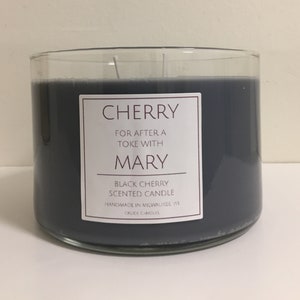 May include: A black cherry scented candle in a clear glass jar with a label that reads "CHERRY FOR AFTER A TOKE WITH MARY BLACK CHERRY SCENTED CANDLE HANDMADE IN MILWAUKEE WI CRUDE CANDLES".