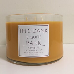 May include: A glass jar candle with a yellow wax and three wicks. The label reads "This dank is quite rank" and "Pumpkin Pie Scented Candle Handmade in Milwaukee, WI Crude Candles".
