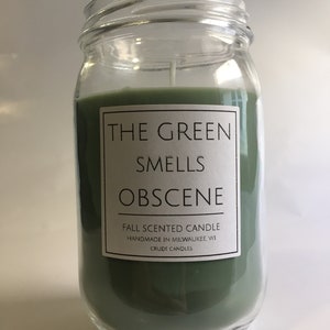 May include: A green glass jar candle with a white label that reads "The Green Smells Obscene" and "Fall Scented Candle Handmade in Milwaukee, WI Crude Candles".
