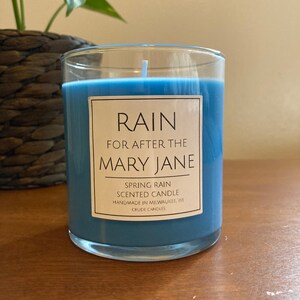 Puede incluir: Un tarro de vela de vidrio azul con una etiqueta blanca que dice "RAIN FOR AFTER THE MARY JANE SPRING RAIN SCENTED CANDLE HANDMADE IN MILWAUKEE WI CRUDE CANDLES".