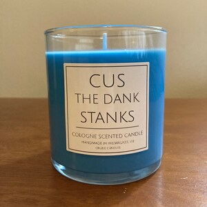 May include: A blue glass candle with a white label that reads "CUS THE DANK STANKS" and "COLOGNE SCENTED CANDLE HANDMADE IN MILWAUKEE, WI CRUDE CANDLES".