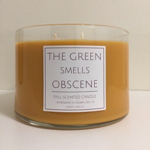Puede incluir: Un tarro de vela de vidrio con una vela amarilla en su interior. La etiqueta dice "The Green Smells Obscene" y "Fall Scented Candle Handmade in Milwaukee, WI Crude Candles".