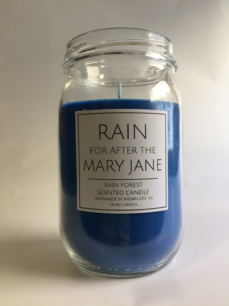 Puede incluir: Una vela perfumada azul en un frasco de vidrio con una etiqueta blanca que dice "RAIN FOR AFTER THE MARY JANE RAIN FOREST SCENTED CANDLE HANDMADE IN MILWAUKEE, WI CRUDE CANDLES".