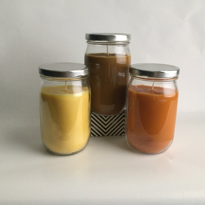 Puede incluir: Tres frascos de vidrio con tapas de metal llenos de velas de diferentes colores. Las velas son amarillas, marrones y naranjas.