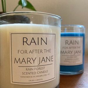 K&ouml;nnte beinhalten: Zwei Glaskerzenbeh&auml;lter mit wei&szlig;em und blauem Wachs. Die Etiketten auf den Beh&auml;ltern lauten "RAIN FOR AFTER THE MARY JANE" und "RAIN FOREST SCENTED CANDLE HANDMADE IN MILWAUKEE WI CRUDE CANDLES".