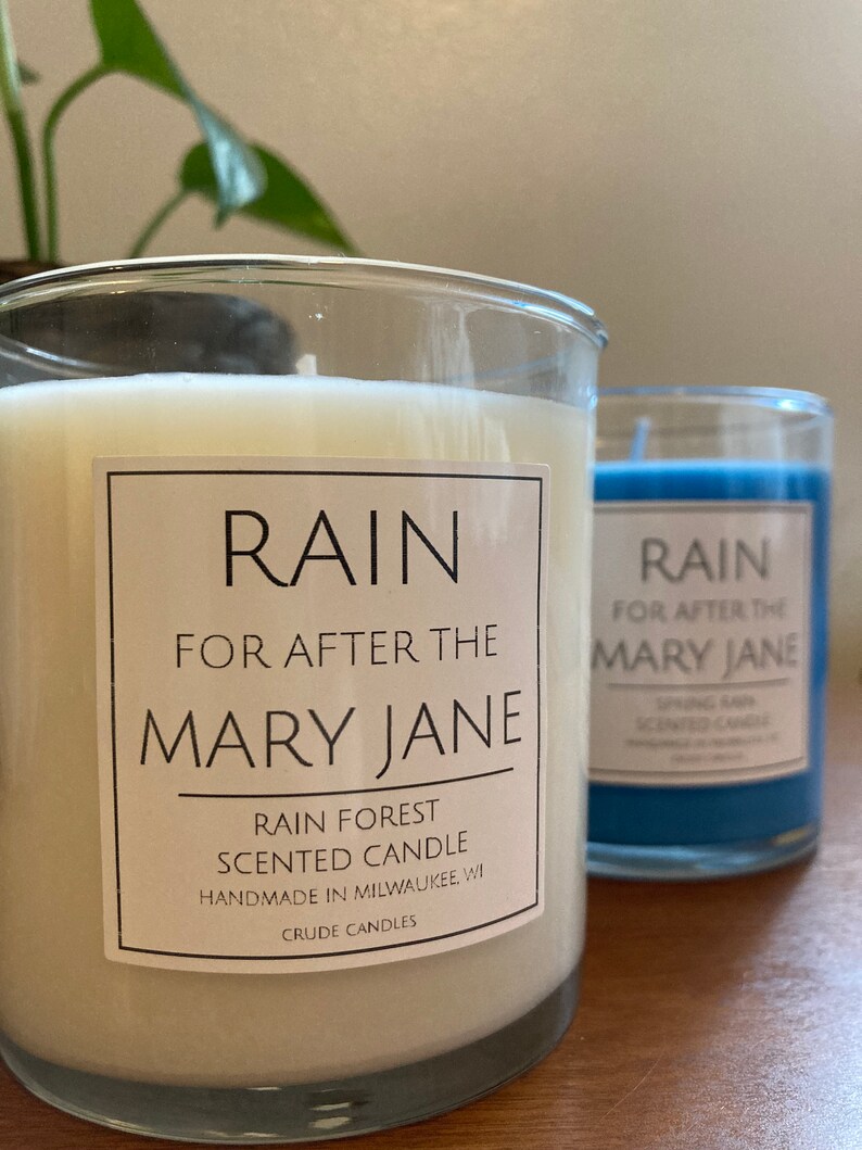 K&ouml;nnte beinhalten: Zwei Glaskerzenbeh&auml;lter mit wei&szlig;em und blauem Wachs. Die Etiketten auf den Beh&auml;ltern lauten "RAIN FOR AFTER THE MARY JANE" und "RAIN FOREST SCENTED CANDLE HANDMADE IN MILWAUKEE WI CRUDE CANDLES".