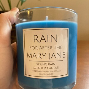 K&ouml;nnte beinhalten: Eine blaue Kerze in einem Glasgef&auml;&szlig; mit einem wei&szlig;en Etikett, auf dem "RAIN FOR AFTER THE MARY JANE SPRING RAIN SCENTED CANDLE HANDMADE IN MILWAUKEE, WI CRUDE CANDLES" steht.