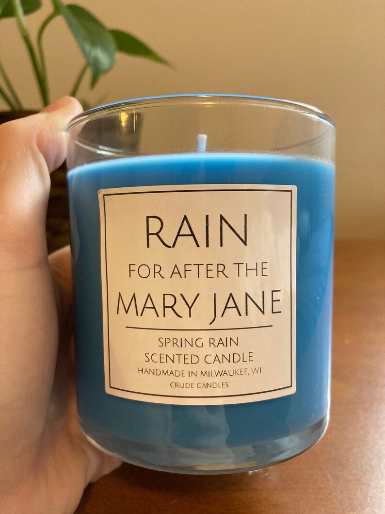 K&ouml;nnte beinhalten: Eine blaue Kerze in einem Glasgef&auml;&szlig; mit einem wei&szlig;en Etikett, auf dem "RAIN FOR AFTER THE MARY JANE SPRING RAIN SCENTED CANDLE HANDMADE IN MILWAUKEE, WI CRUDE CANDLES" steht.