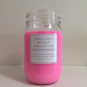 Puede incluir: Una vela de vidrio rosa en un frasco con una etiqueta blanca que dice "Huele a chico, pero sin la decepci&oacute;n." La etiqueta tambi&eacute;n dice "Vela perfumada con colonia" y "Hecho a mano en Milwaukee, WI".