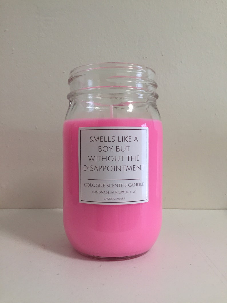 Puede incluir: Una vela de vidrio rosa en un frasco con una etiqueta blanca que dice "Huele a chico, pero sin la decepci&oacute;n." La etiqueta tambi&eacute;n dice "Vela perfumada con colonia" y "Hecho a mano en Milwaukee, WI".