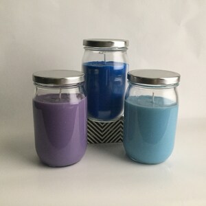 Puede incluir: Tres tarros de vidrio para velas con tapas plateadas. Las velas son moradas, azules y azul claro.