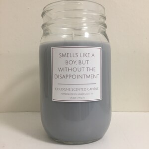 Puede incluir: Una vela de vidrio gris en un frasco con una etiqueta blanca que dice "Huele como un chico, pero sin la decepci&oacute;n." La etiqueta tambi&eacute;n dice "Vela perfumada con colonia" y "Hecho a mano en Milwaukee WI".